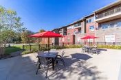 Sunlit outdoor patio with round tables, chairs, and red umbrellas beside a multi-story brick senior living building and landscaped lawn under a blue sky.