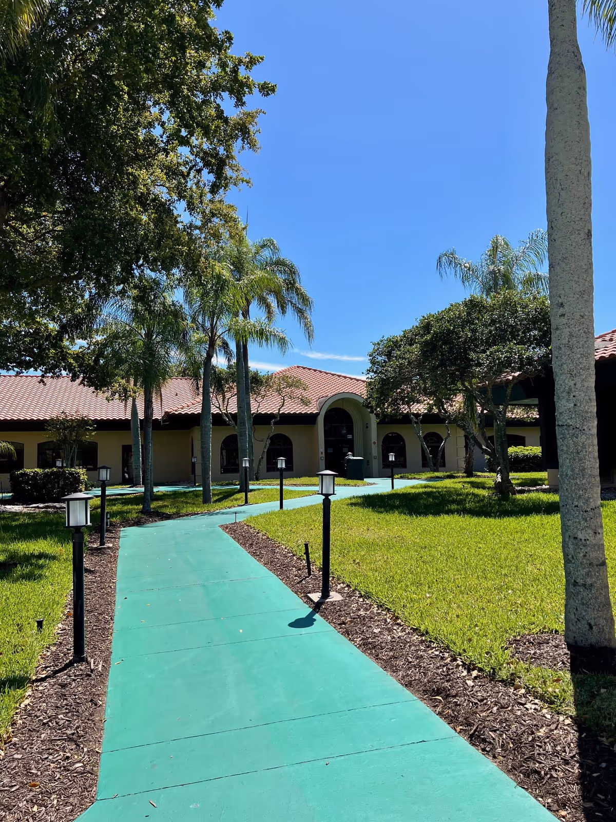 A sunny outdoor pathway painted green leads through a landscaped garden with palm trees and other greenery towards a single-story building with a red-tiled roof and arched windows and doorways under a clear blue sky.