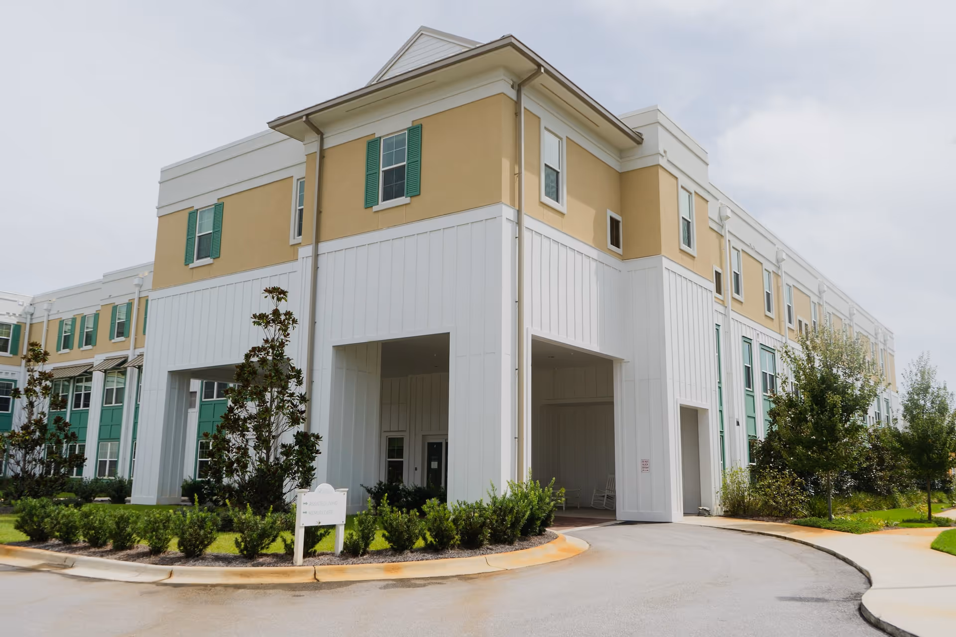 Exterior view of a multi-story senior living facility building with beige and white walls, green window shutters, landscaped bushes, and a driveway leading to a covered entrance.
