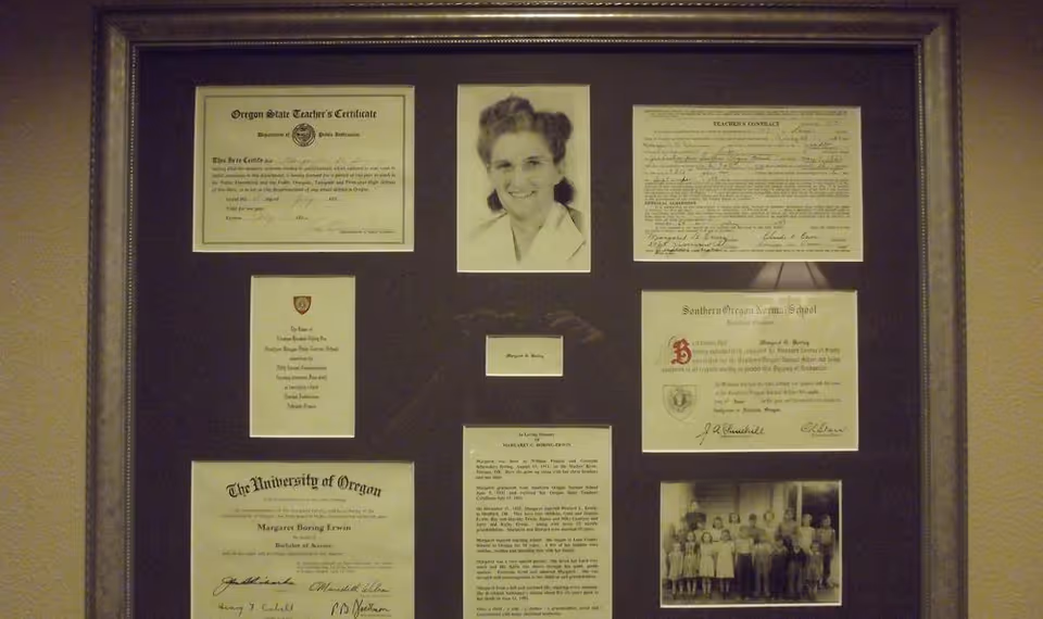 Framed display of diplomas, certificates and black-and-white photos hung on a beige wall.