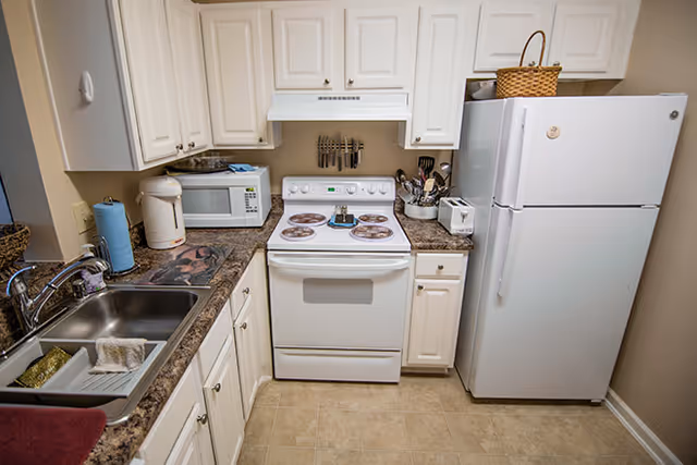 A compact kitchen with white cabinets and appliances including a refrigerator, stove, microwave, and sink. The countertops are brown with a speckled pattern, and there are various kitchen items such as a paper towel holder, knife block, and utensil holder. The floor is tiled in a light beige color.