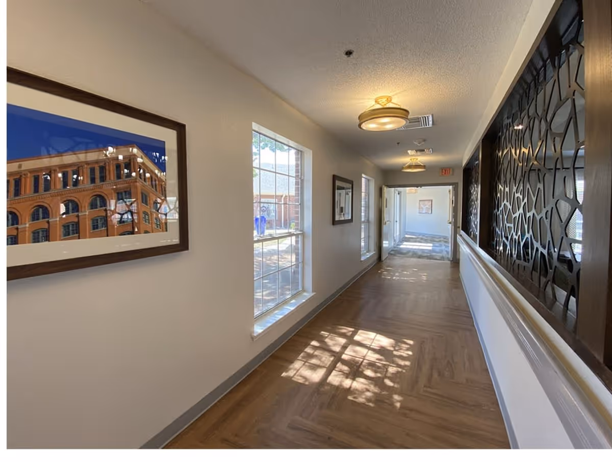 A well-lit hallway with wooden flooring and white walls. On the left wall, there are two windows letting in natural light and a framed picture of a building. On the right side, there is a decorative dark panel with an abstract pattern. Ceiling lights illuminate the corridor, and an exit sign is visible at the far end.