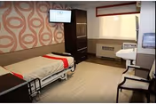 A rehabilitation and nursing center room featuring a single hospital bed with white and red bedding, a dark wooden cabinet, a wall-mounted television, a window with a beige roller shade, a heating unit below the window, a sink with a mirror, and two chairs with wooden frames and cushioned seats.