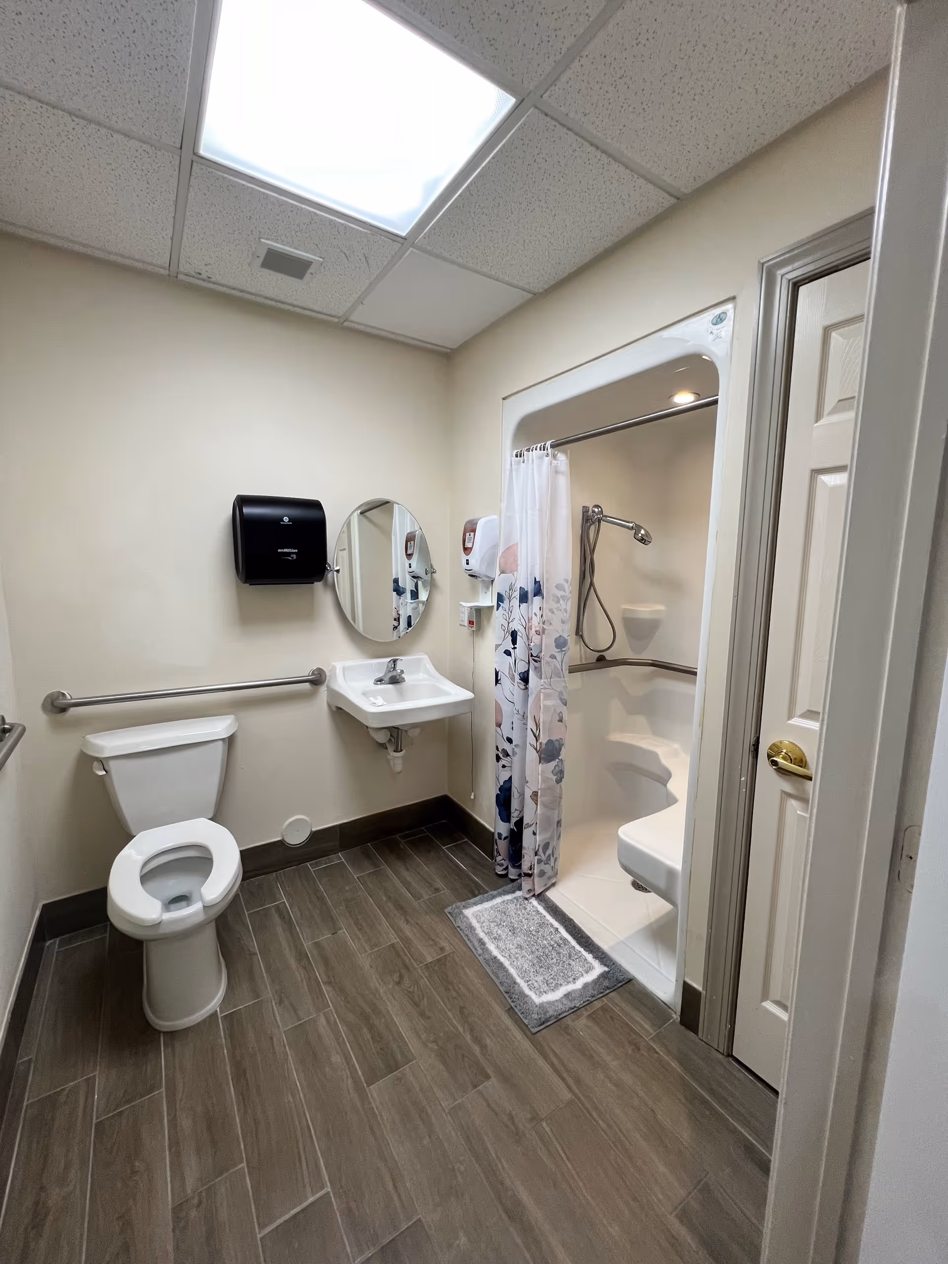 A clean, accessible bathroom with a toilet, wall-mounted sink, round mirror, paper towel dispenser, and a shower with a floral shower curtain and built-in seat. The floor has wood-look tiles and the walls are painted light beige.