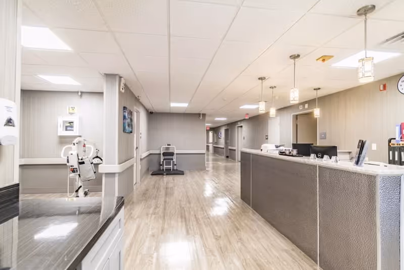 A clean and well-lit nursing facility hallway with a reception desk on the right side, pendant lights hanging from the ceiling, and medical equipment visible in a small alcove on the left. The floor is light wood, and the walls are painted in neutral tones.