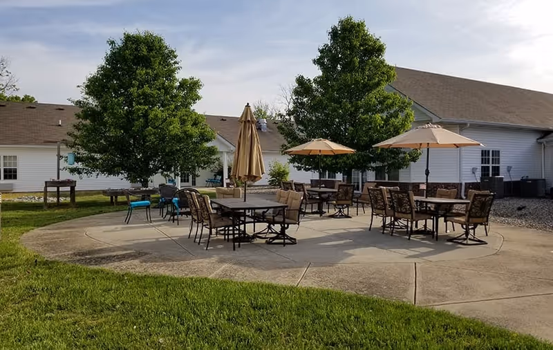 Outdoor patio area with several tables and chairs, some shaded by large umbrellas, surrounded by green grass and trees, adjacent to a white building with a brown roof under a partly cloudy sky.
