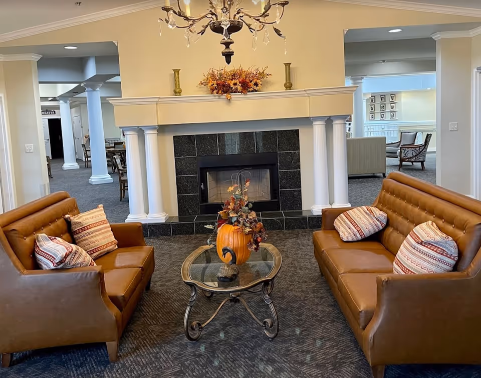 A cozy living room area with two brown leather sofas facing each other, each with patterned throw pillows. Between them is a glass-top coffee table with a decorative pumpkin and autumn-themed floral arrangement. Behind the table is a fireplace with a black tile surround and white mantel decorated with two green vases and a fall floral arrangement. The room has beige walls, carpeted flooring, and a chandelier hanging from the ceiling. In the background, there are additional seating areas and white columns.