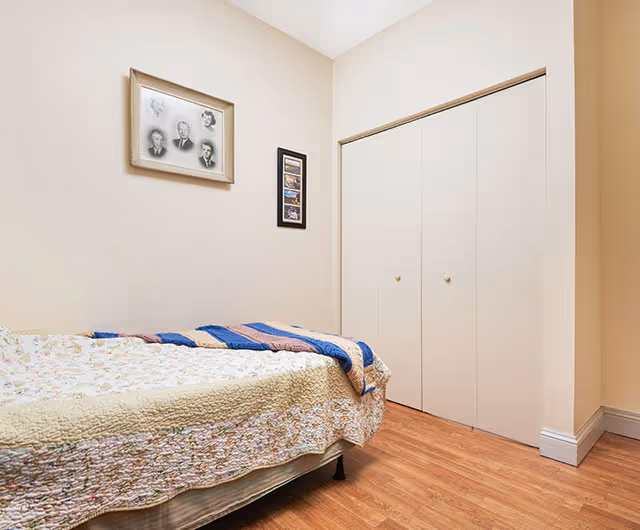 Small bedroom with a single bed covered by a patchwork quilt, framed pictures on beige walls, and a closed double-door closet.