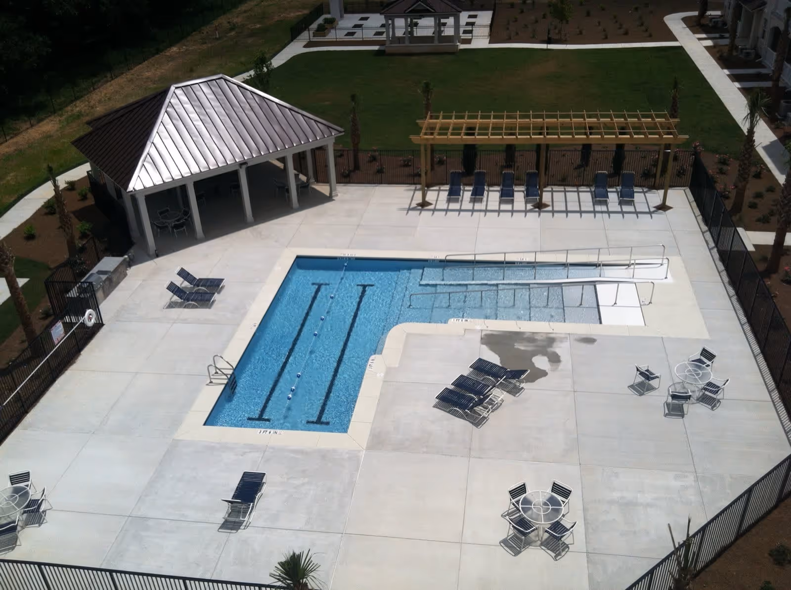 Aerial view of an outdoor swimming pool area at Fairfield Manor featuring a rectangular pool with lane markers, a smaller pool with a ramp for accessibility, several lounge chairs, round tables with chairs, a covered pavilion with seating, and a pergola with additional lounge chairs. The area is surrounded by a fence and landscaped with grass and small plants.
