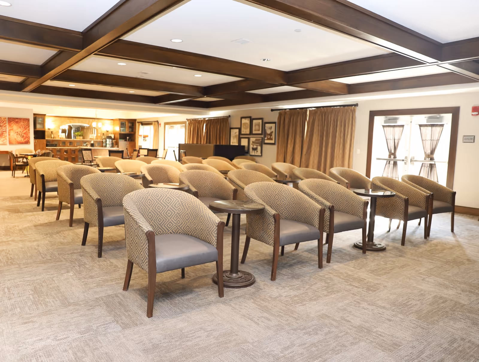 A spacious room with multiple rows of patterned armchairs arranged in pairs, each pair separated by small round tables. The room features a coffered ceiling with wooden beams, beige carpeted floor, and large windows with brown curtains. In the background, there is a counter area with chairs and framed artwork on the walls.