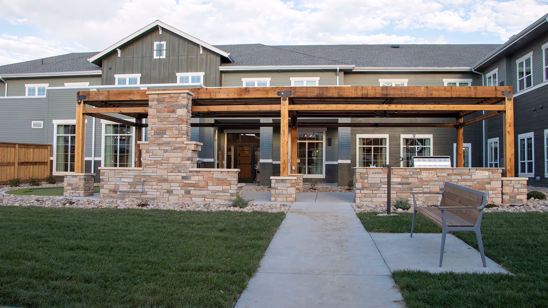 Front exterior of a senior living building featuring a covered wooden pergola with stone columns, a central walkway, bench, and lawn.