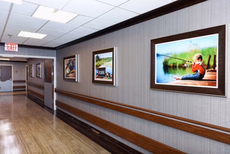 A well-lit hallway with wooden handrails on both sides and framed pictures hanging on the right wall. The floor is polished wood, and there is an exit sign visible at the end of the corridor.