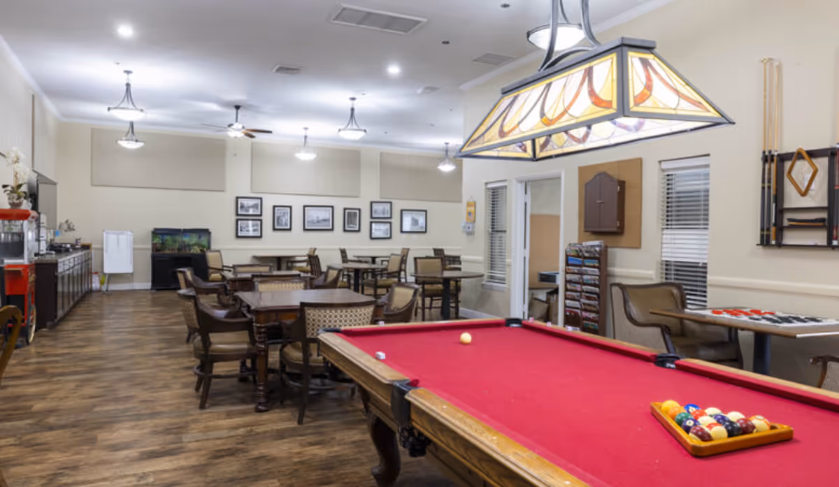 Interior view of a recreational room in a retirement community featuring a red felt pool table with balls arranged on it, several tables and chairs for seating, framed pictures on the wall, a fish tank, and a popcorn machine. The room has wooden flooring and ceiling lights.
