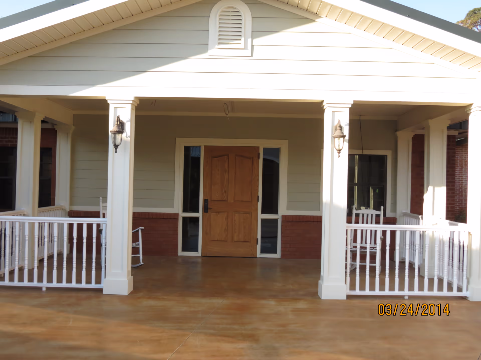 Front porch of a building with a wooden door in the center, flanked by two windows. The porch has white railings and columns, two wall-mounted lantern-style lights, and two white rocking chairs on either side. The ground is a polished concrete surface.