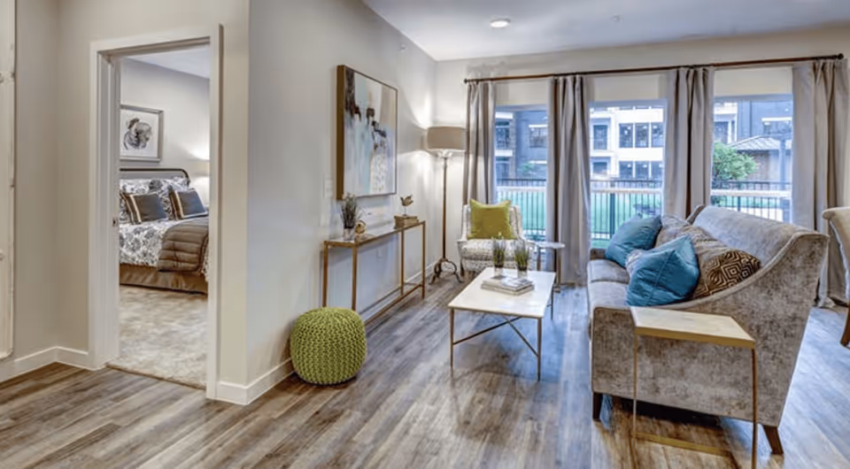 A cozy living room with a gray sofa adorned with blue and patterned pillows, a white coffee table with decorative items, an armchair with a green pillow, and large windows with beige curtains overlooking an outdoor area. To the left, there is a hallway leading to a bedroom with a bed covered in patterned bedding and pillows. The room has wood flooring and neutral-colored walls with a piece of artwork and a floor lamp.