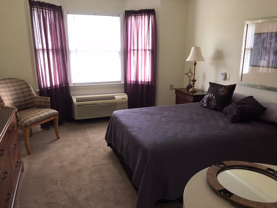 A bedroom with a bed covered in a purple quilt and matching pillows, a wooden nightstand with a lamp and decorative cross, a window with purple curtains, a plaid upholstered chair, a wooden dresser, and a round table with a decorative mirror on top.
