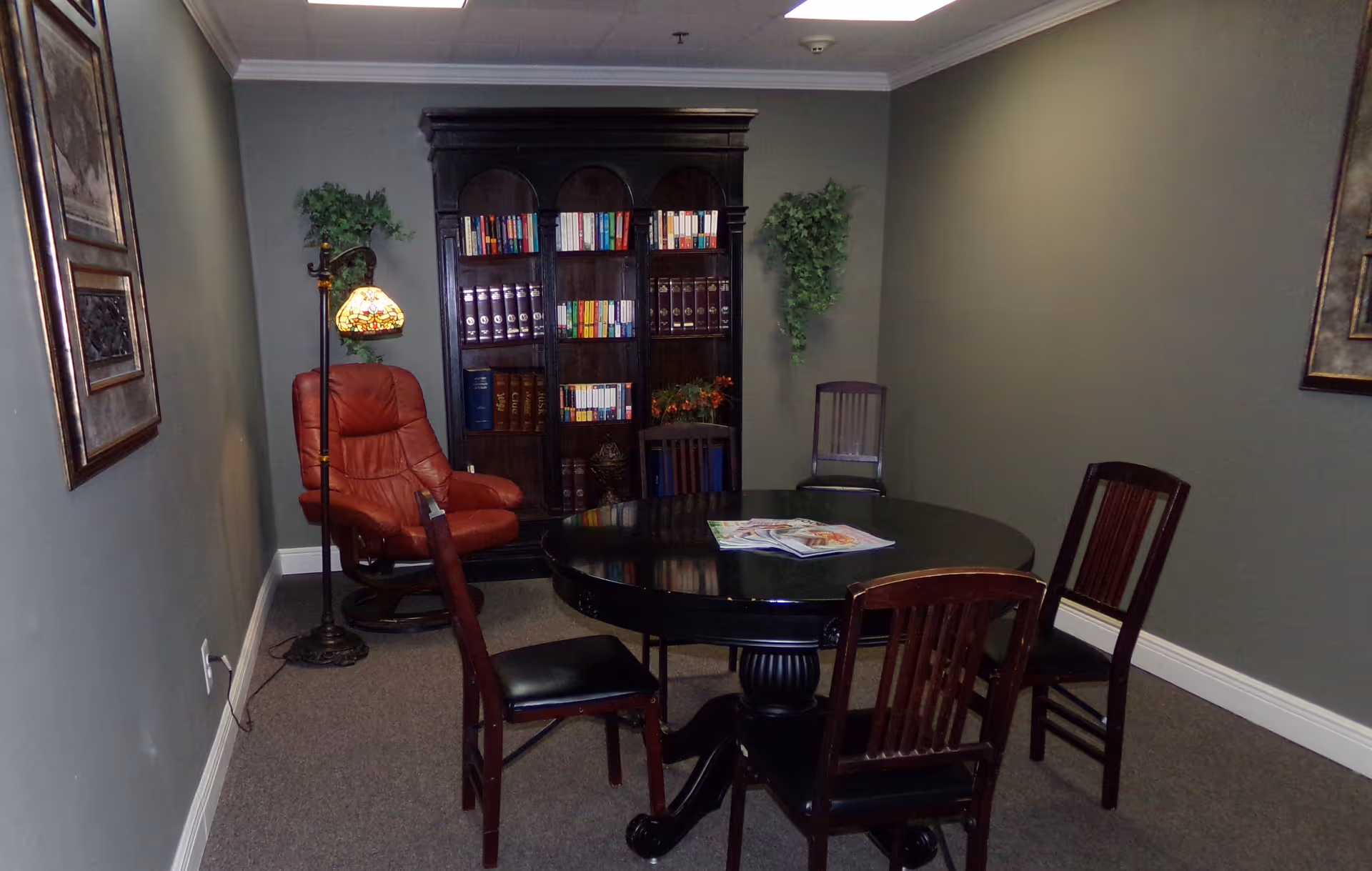Small interior sitting room with a round black table and chairs, a bookshelf, a red recliner and a standing lamp.