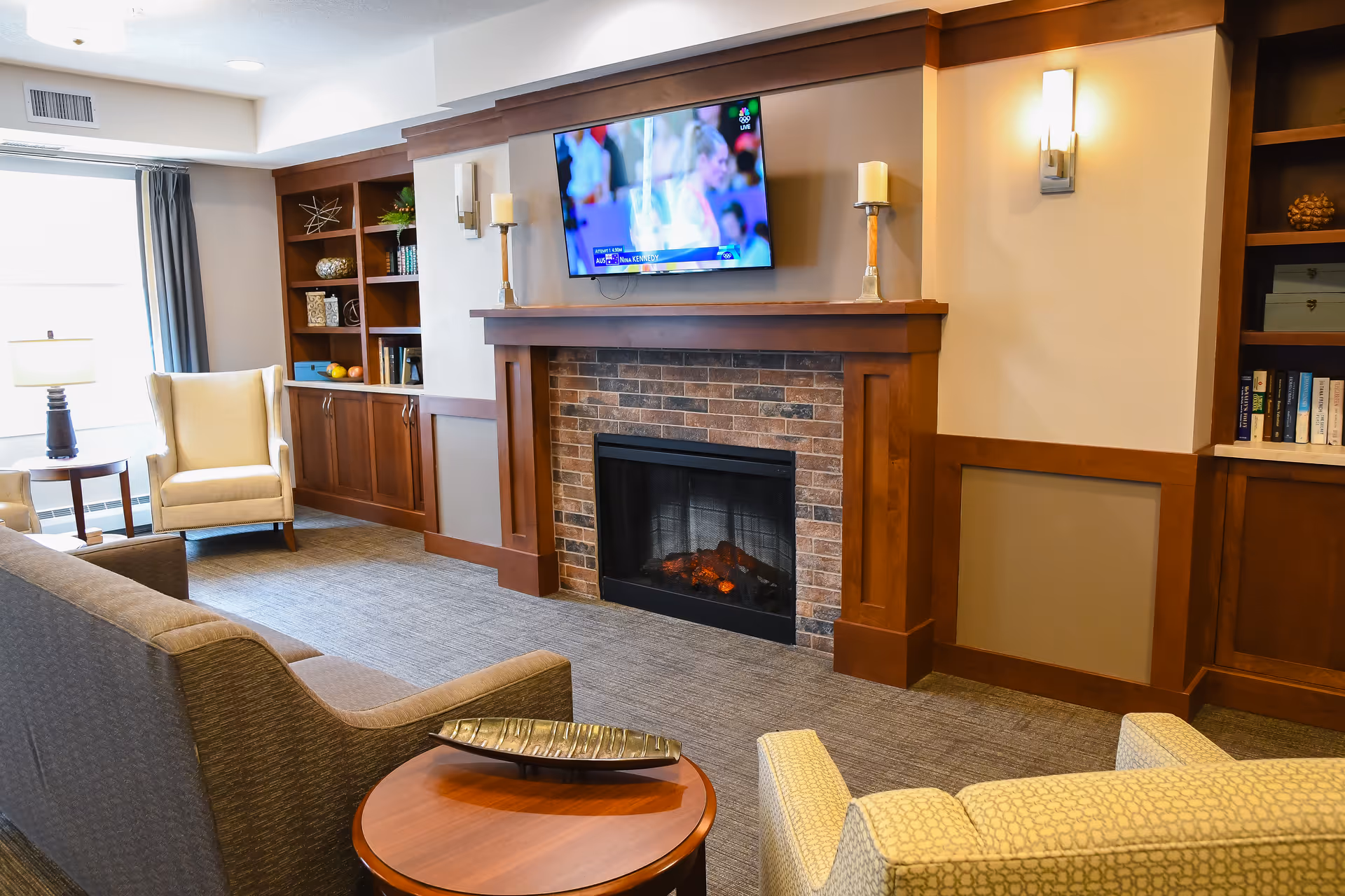 A cozy living room area featuring a brick fireplace with a wooden mantle, a wall-mounted TV above it, built-in wooden shelves on either side filled with books and decorative items, a beige armchair near a window with curtains, a gray sofa, and a round wooden coffee table with a decorative tray.