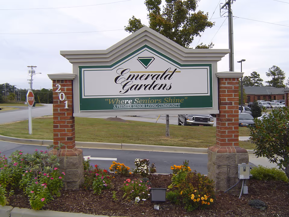 Outdoor sign for Emerald Gardens senior living community with the tagline 'Where Seniors Shine' surrounded by flowers and greenery, with a parking lot and buildings in the background.