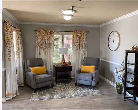 A cozy living room area with two gray armchairs each with a yellow cushion, positioned on a patterned rug. Behind the chairs is a window with white and yellow floral curtains and a small table with a lamp and autumn decorations. A large round clock hangs on the wall to the right, and a tall wooden shelf with decorative items stands in the corner.