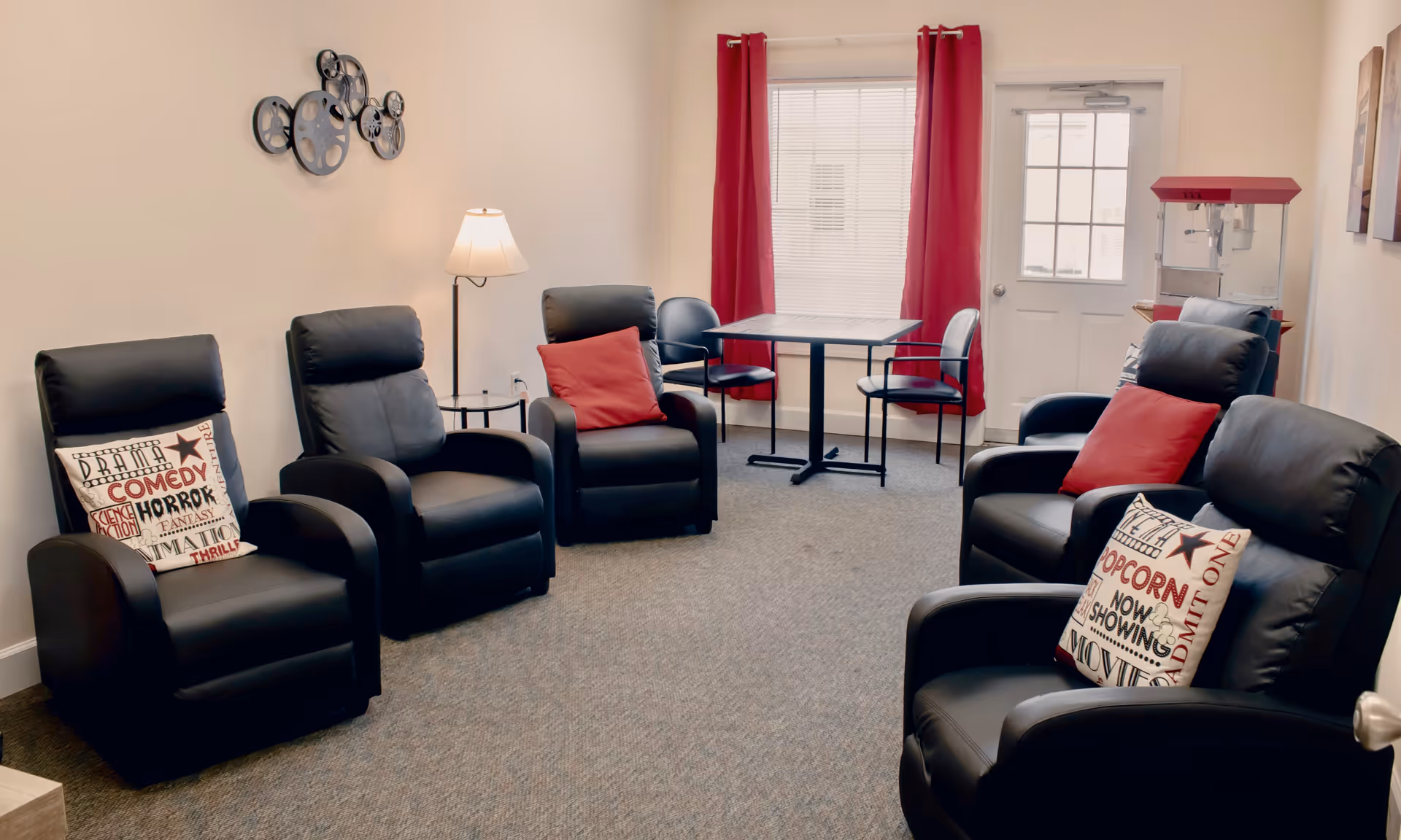 A cozy common room with six black leather recliners arranged in a semi-circle, each with decorative pillows featuring movie-themed text. There is a floor lamp and a small round table between two chairs on the left side. At the back of the room, there is a small square table with four chairs, red curtains on the window, and a door with window panes. A red popcorn machine is placed near the door on the right side. The walls are light-colored and the floor is carpeted.