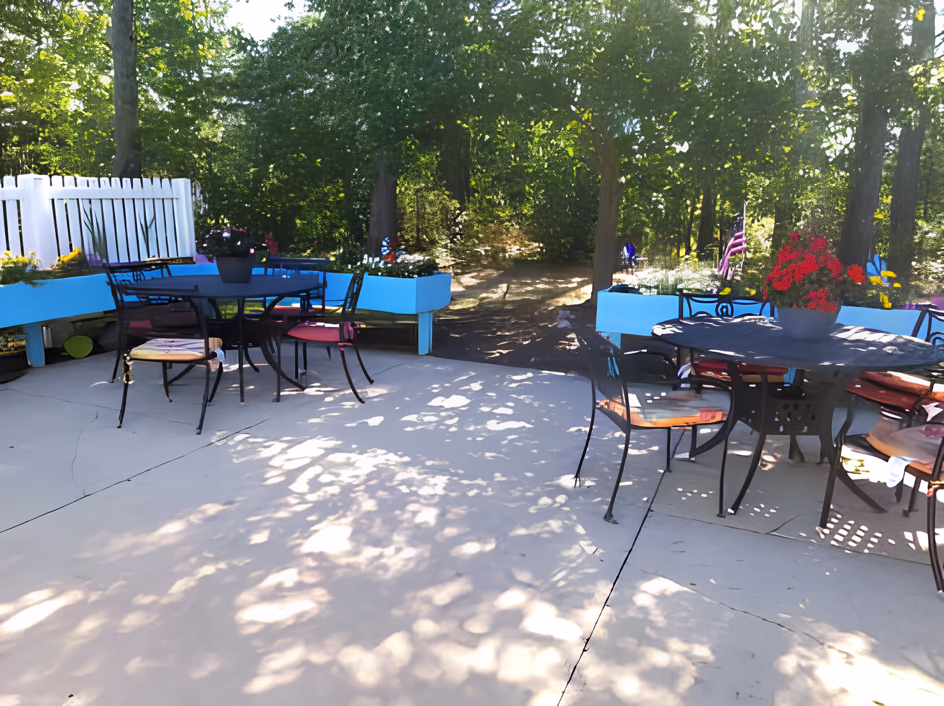 Outdoor patio with round tables and chairs, bright blue planter boxes and potted flowers under trees and dappled sunlight.