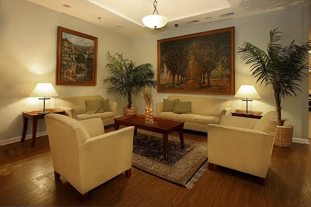 A cozy living room with beige upholstered sofas and armchairs arranged around a wooden coffee table on a patterned rug. Two large framed paintings hang on the walls, and two potted plants are placed in the corners. The room is softly lit by table lamps on side tables and a ceiling light fixture.