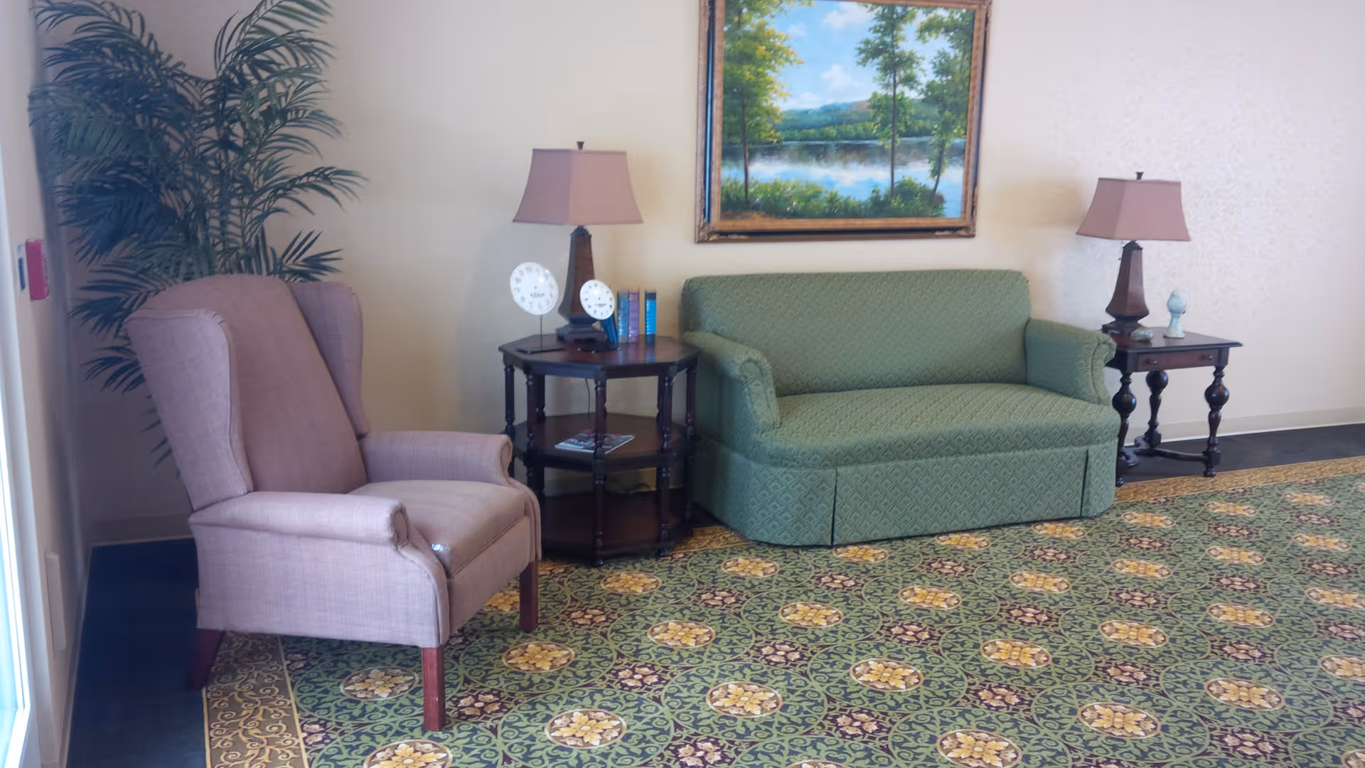 A cozy sitting area with a green loveseat, mauve wingback chair, side tables with lamps, a potted plant and a landscape painting on patterned carpet.