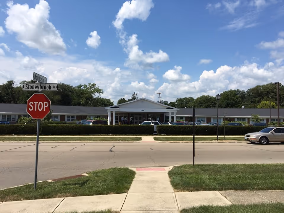 Exterior view of The Trinity Community at Fairborn building on a sunny day with a partly cloudy sky. The building is a single-story structure with a white entrance canopy and several cars parked in front. A stop sign and street signs for Monticello and Stoneybrook Trail are visible in the foreground along a paved road with sidewalks and grass.