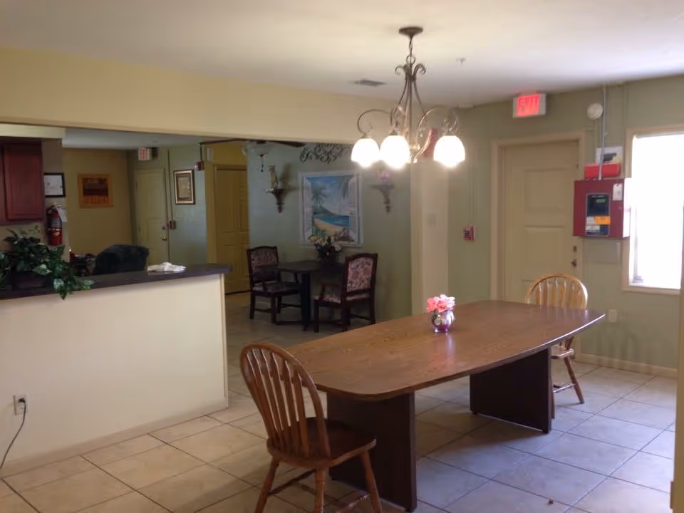 Communal dining area with a long wooden table, chairs, a hanging chandelier, and a small seating nook in the background.