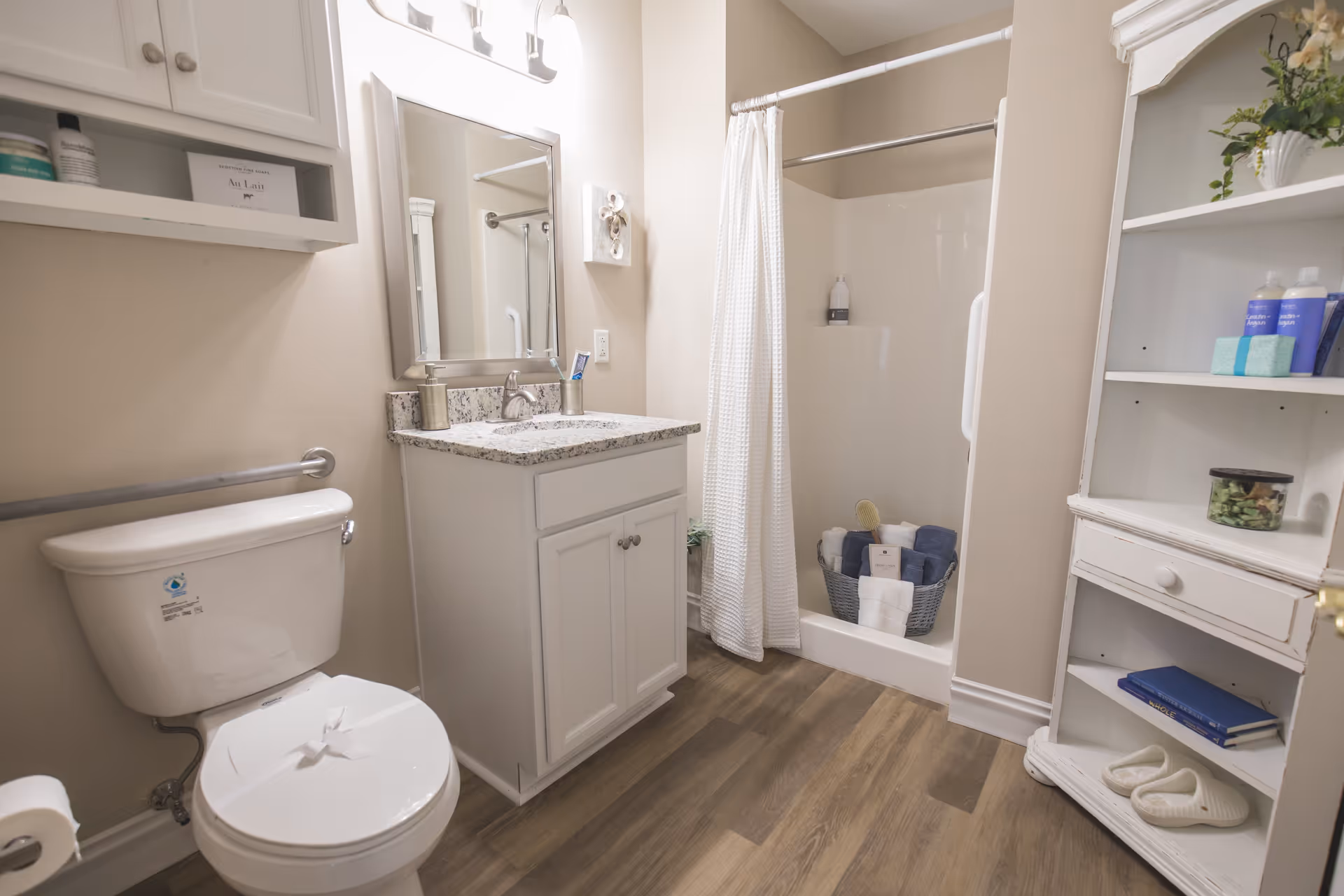 A clean and well-lit bathroom featuring a white toilet with a safety grab bar, a white vanity with a granite countertop and a mirror above it, a shower with a white curtain and a basket of towels inside, and a white shelving unit holding toiletries, books, and slippers. The floor is wood-style laminate.
