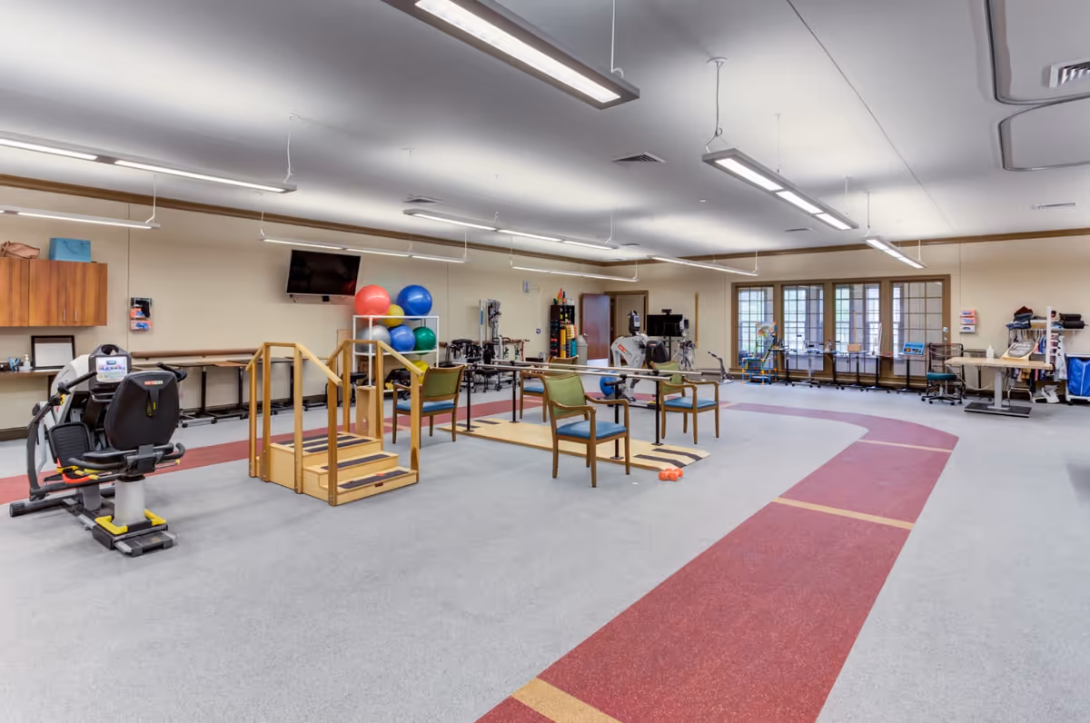A spacious rehabilitation and exercise room with various physical therapy equipment including parallel bars, exercise balls, a stationary bike, and chairs. The room has a red and gray floor with a walking track, large windows letting in natural light, and ceiling-mounted fluorescent lights.