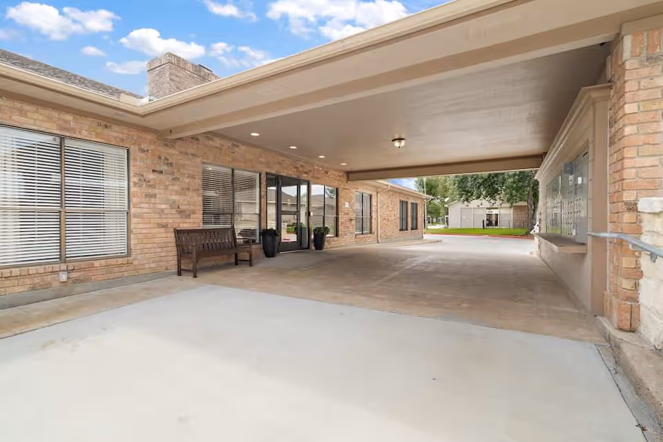 Covered entrance area of a brick building with a bench, large windows with blinds, double glass doors, and a view of a green lawn and trees in the background under a partly cloudy sky.