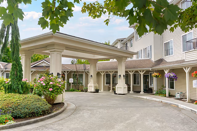 Entrance area of a senior living facility with a covered drop-off zone, surrounded by well-maintained landscaping including bushes and hanging flower baskets. The building exterior is light-colored with multiple windows and a three-story structure.
