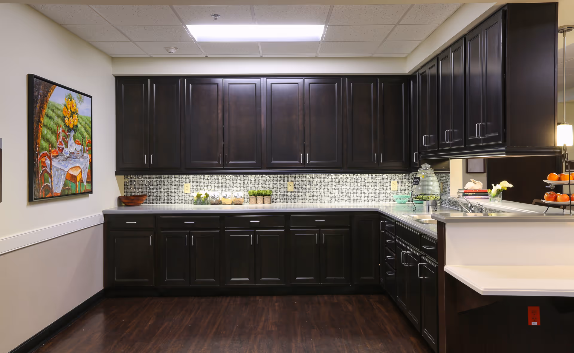 A modern kitchen with dark wooden cabinets, a mosaic tile backsplash, and a white countertop. The kitchen features a sink, a tiered fruit stand with oranges, and decorative items including small plants and jars. A colorful painting of a table set outdoors with flowers hangs on the left wall.
