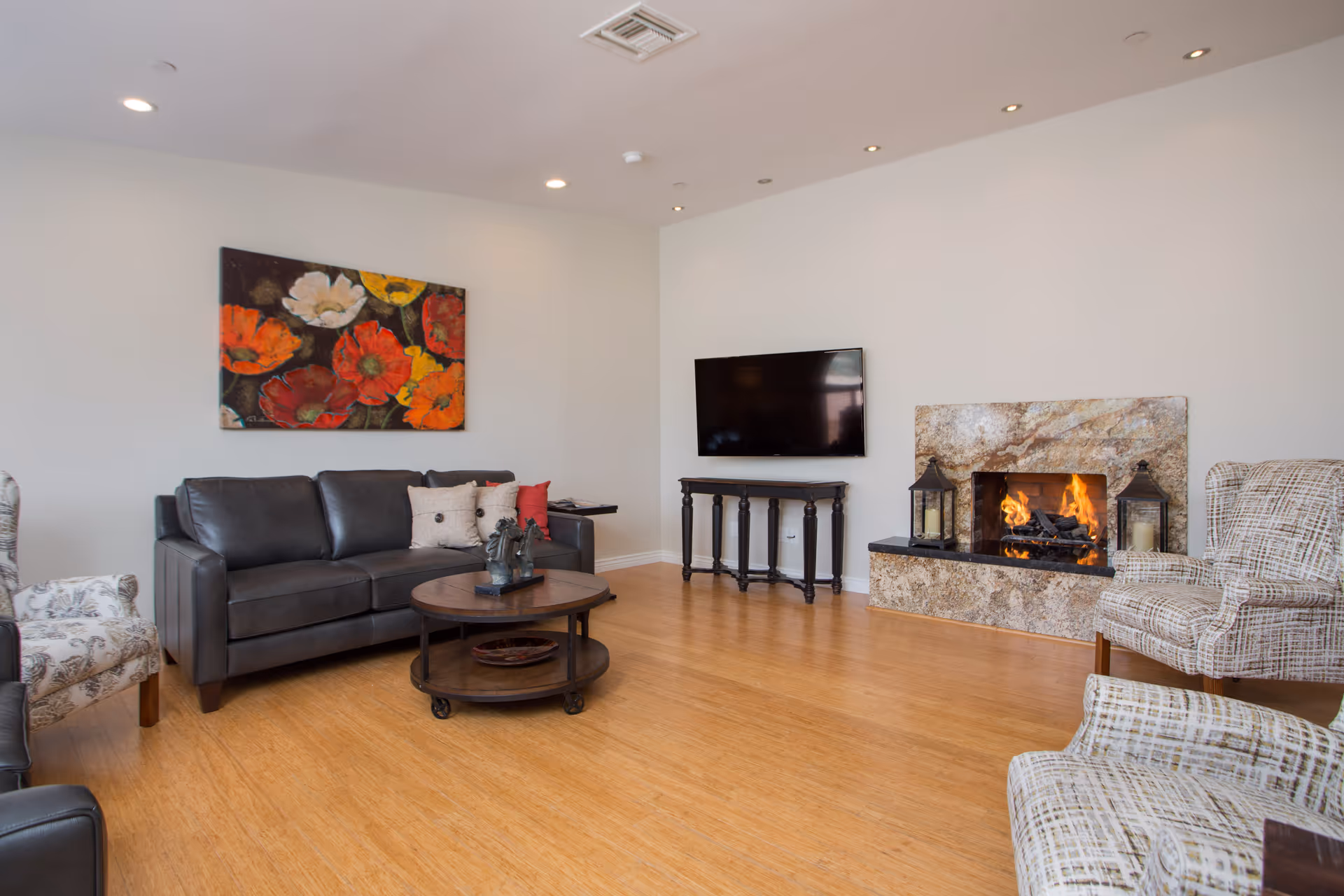 A cozy living room with a dark leather sofa adorned with pillows, two patterned armchairs, a round wooden coffee table with decorative items, a wall-mounted flat-screen TV, and a fireplace with a stone surround and two lanterns on either side. A colorful floral painting hangs on the wall above the sofa.