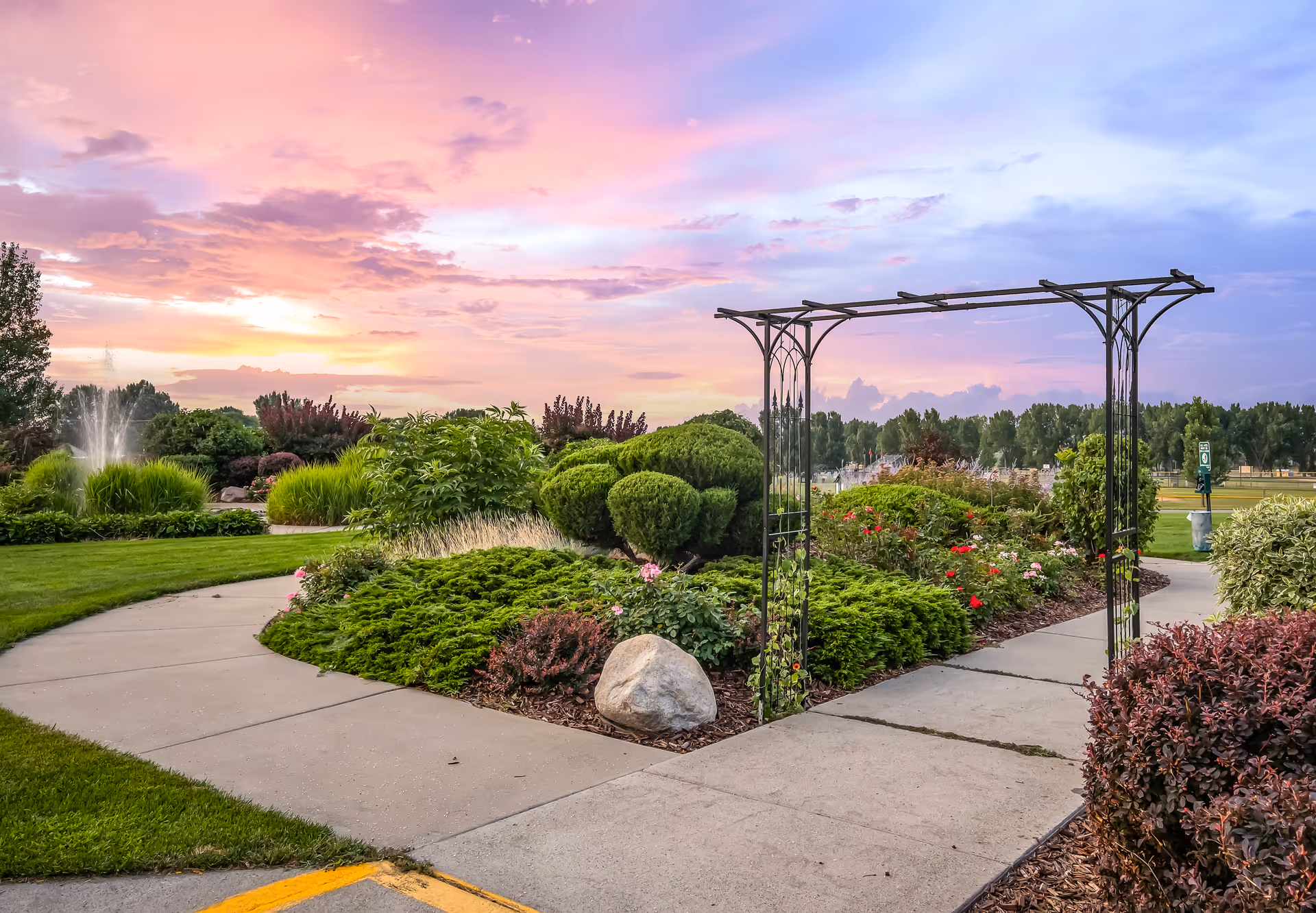 A landscaped outdoor garden area at sunset with a paved walkway, green bushes, flowering plants, a metal archway, and a water fountain in the background under a colorful sky.