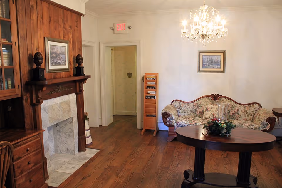 Traditional sitting room with a floral upholstered sofa, wooden fireplace, round table and chandelier.