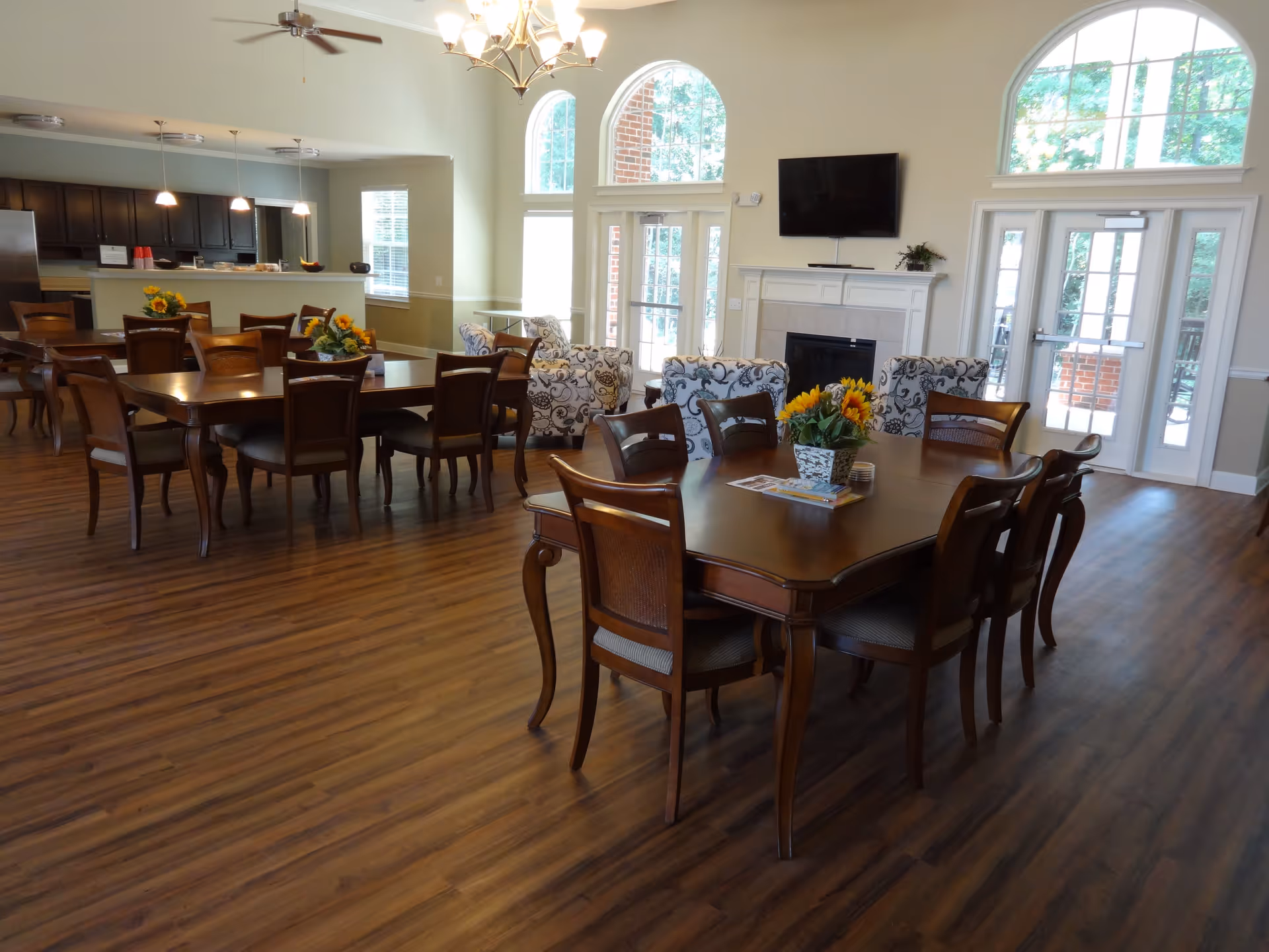 A spacious senior living common area with wooden floors, multiple wooden dining tables and chairs, a kitchen area with dark cabinets and hanging lights, a fireplace with a mounted TV above it, and large arched windows letting in natural light. There are also patterned armchairs near the fireplace and a door leading outside.