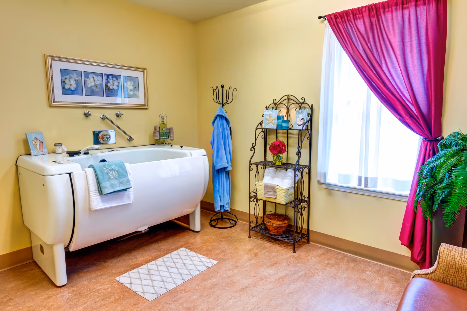 A bright bathroom with a large white walk-in bathtub against a yellow wall. Above the tub is a framed picture of white flowers. A blue towel and a white towel hang on the tub. To the right of the tub is a black metal rack holding rolled white towels, decorative items, and a vase with red flowers. A blue robe hangs on a black coat rack near the corner. A window with sheer white curtains and bright pink drapes is on the right wall, next to a green potted plant and a wicker chair.
