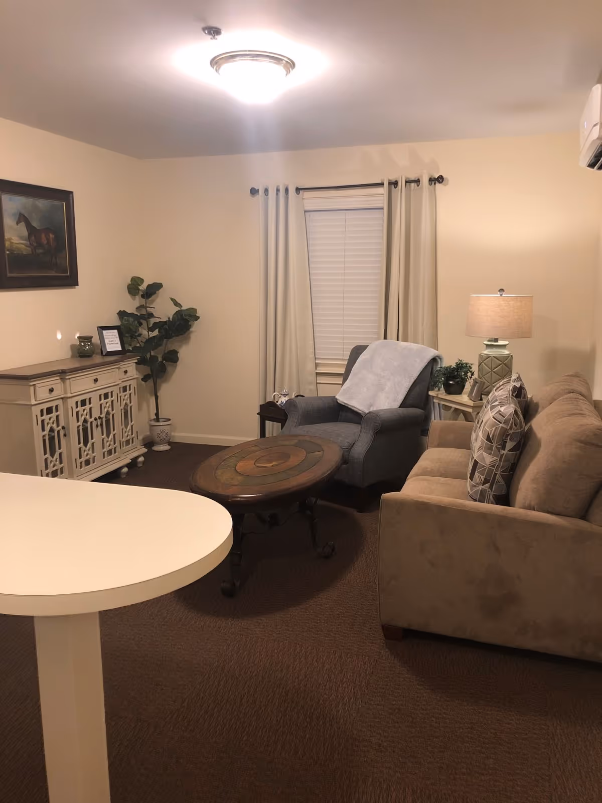 A cozy living room with a beige sofa adorned with patterned pillows, a gray armchair with a light gray throw blanket, a round wooden coffee table, a white cabinet with decorative items, a potted plant, a table lamp on a side table, and a window with closed blinds and beige curtains.