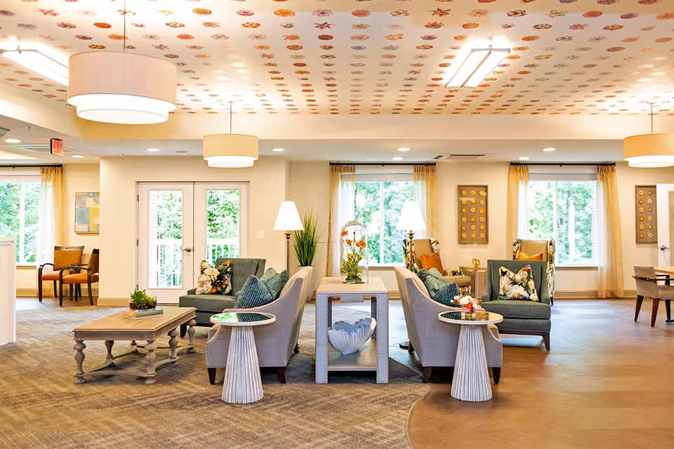 A bright and spacious living room area in a senior living facility with comfortable armchairs and sofas arranged around coffee and side tables. The ceiling is decorated with a pattern of colorful leaves, and large windows with sheer curtains allow natural light to fill the room. There are lamps and plants placed on tables, creating a cozy and inviting atmosphere.