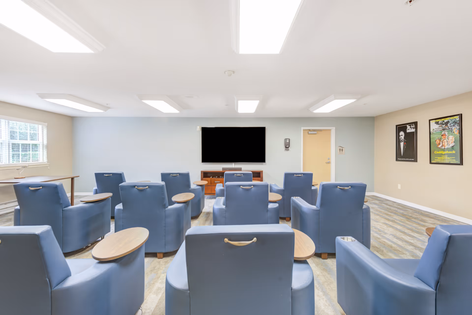 A bright room with multiple blue armchairs arranged in rows facing a large flat-screen TV mounted on a light blue wall. The room has a window on the left side, two framed posters on the right wall, and a door next to the TV. The floor has a patterned carpet, and the ceiling has several rectangular fluorescent lights.