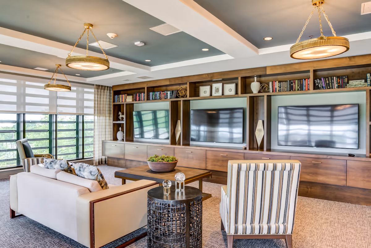A bright and modern living room with large windows covered by sheer blinds, two beige sofas with patterned pillows, a striped armchair, a wooden coffee table with a decorative bowl, and a built-in wooden entertainment center featuring three large flat-screen TVs and shelves with books and decorative items. Two round pendant lights hang from the ceiling.