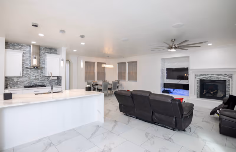 Open-plan modern living room and kitchen with white marble floors, a large island, dining table, dark leather recliner sofa, wall-mounted TV and fireplace.