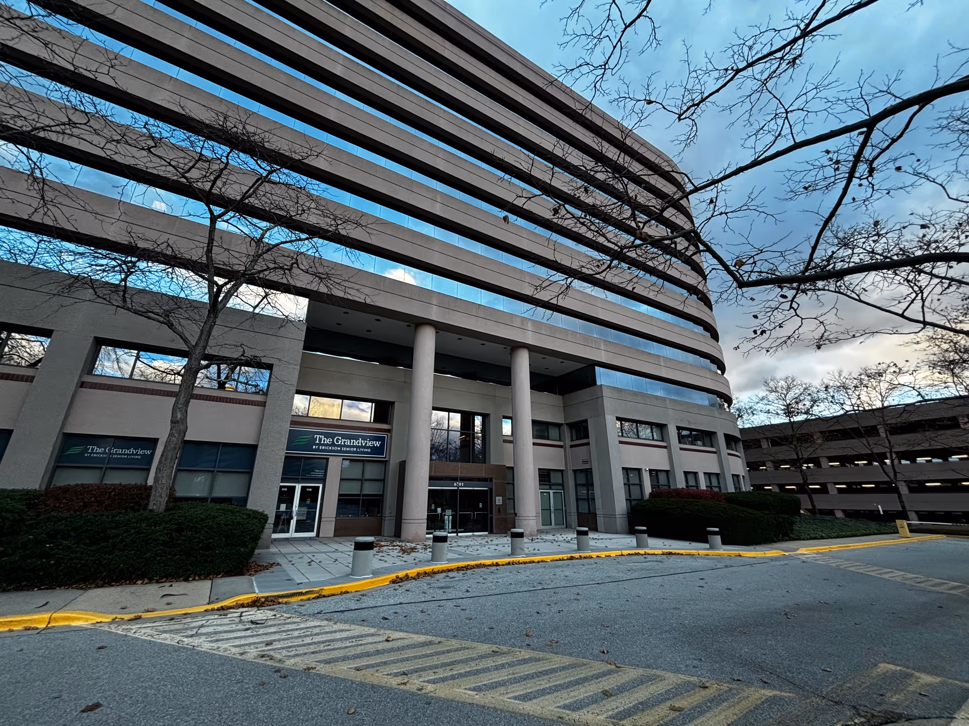 Exterior view of a multi-story building with large windows and columns at the entrance. The building has signs that read 'The Grandview by Erickson Senior Living'. Leafless trees and bushes are visible around the entrance, and a parking structure is seen to the right.