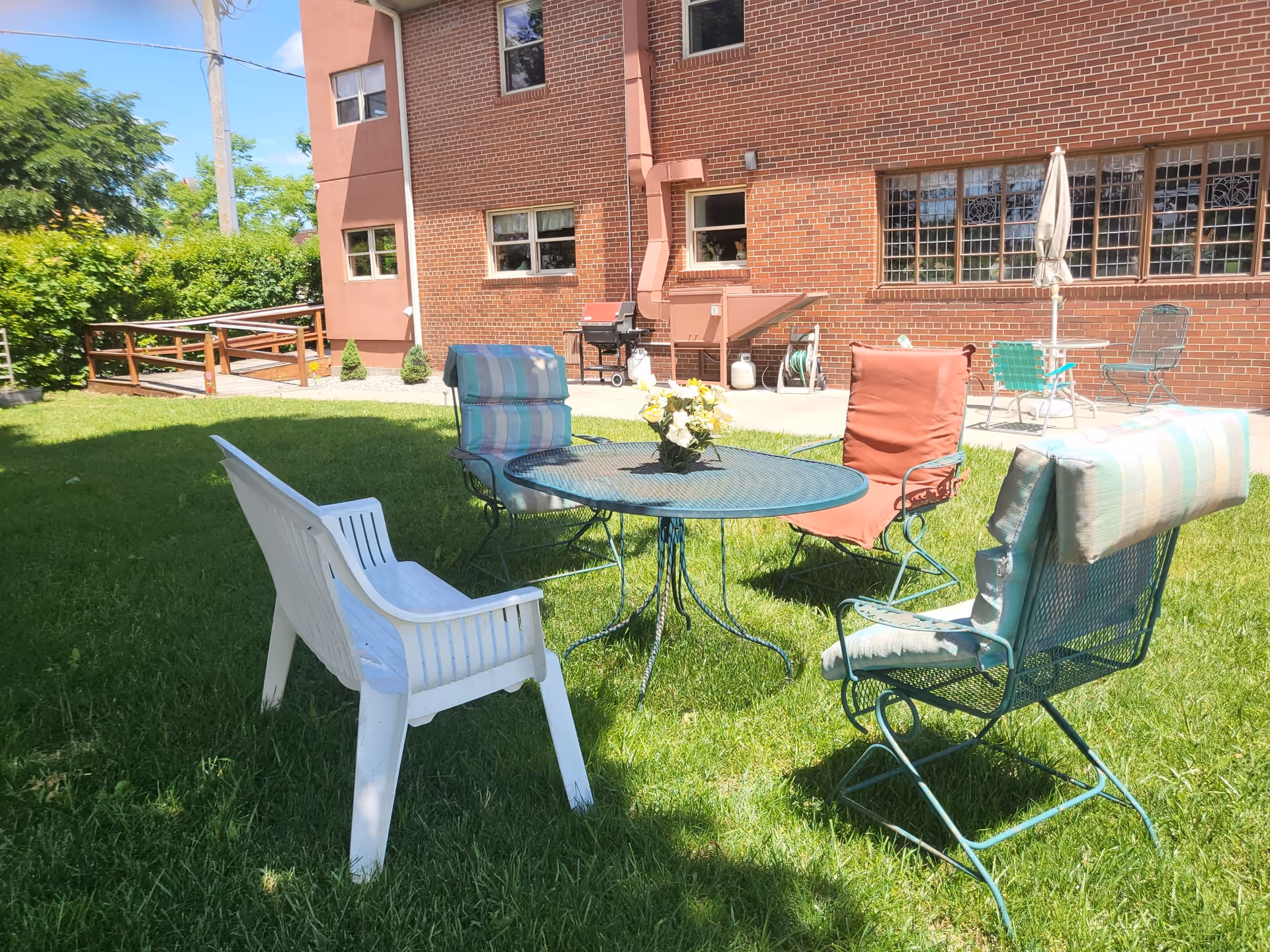 Outdoor patio area with a round metal table and four chairs on green grass. Three chairs have cushions, and one is a white plastic chair. A small flower arrangement is on the table. In the background, there is a brick building with windows, a grill, and another small table with chairs and an umbrella.