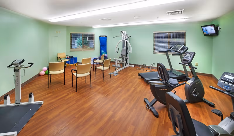 A fitness room with light green walls and wood flooring featuring exercise equipment including a treadmill, stationary bike, weight machine, and chairs arranged in a semi-circle. Two windows with blinds and a small wall-mounted TV are visible.