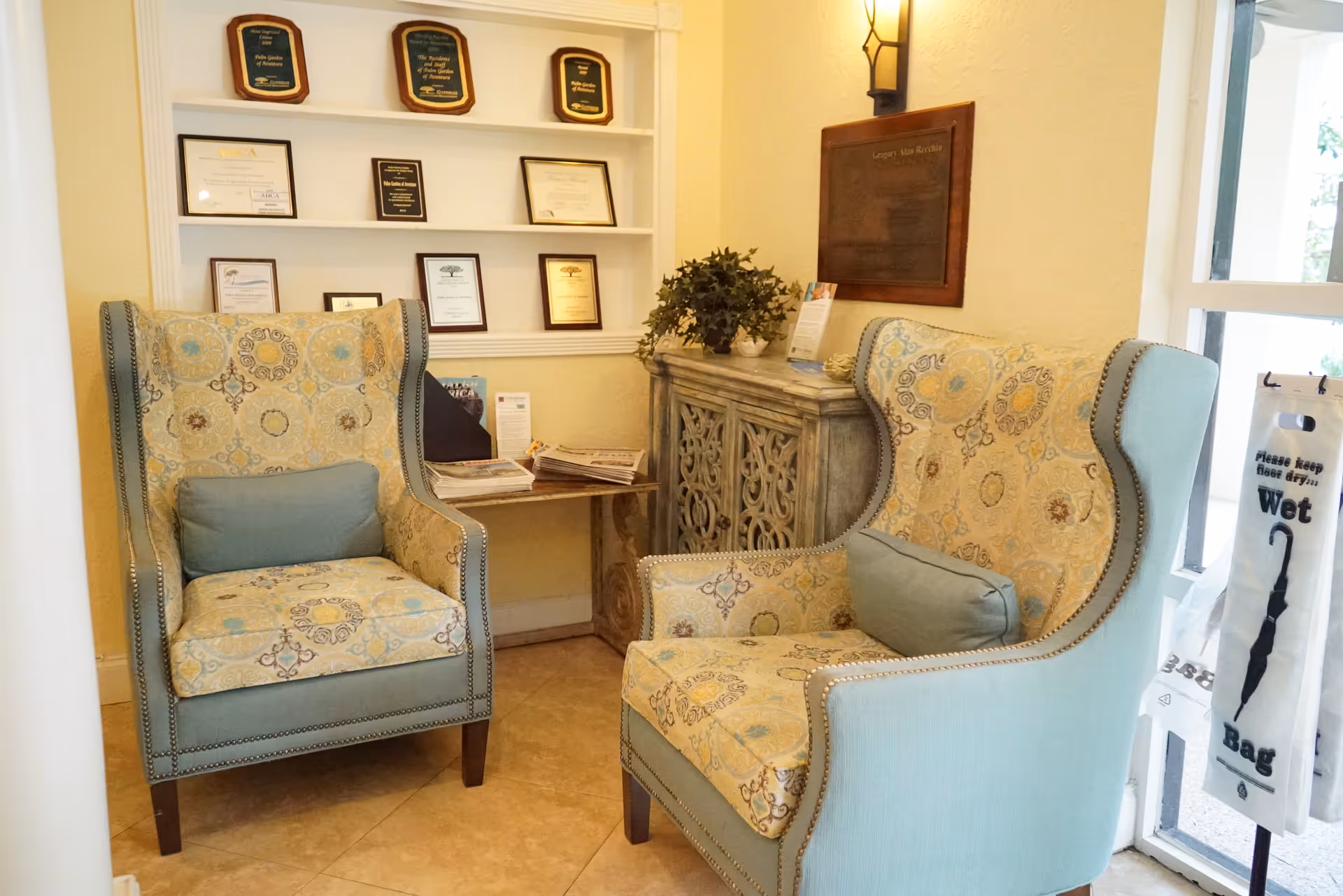 Two patterned wingback chairs with a small table, decorative cabinet, and plaques on the wall in a facility lobby.