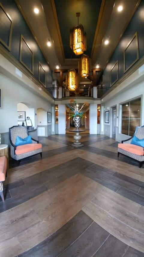 Spacious and elegant interior lobby area with high ceilings, three large hanging lantern-style lights, patterned wood flooring, and a central decorative pedestal with a floral arrangement. Two cushioned armchairs with blue pillows are positioned on either side, and there are windows and doorways along the walls.